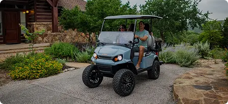 Sturdy cyan golf cart with large wheels driven by a person wearing shorts and a gray shirt, slightly elevated compared to a standard golf cart