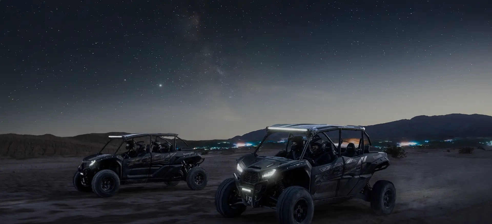 Two black adventure cars driving over desert dunes at night under a sky full of stars