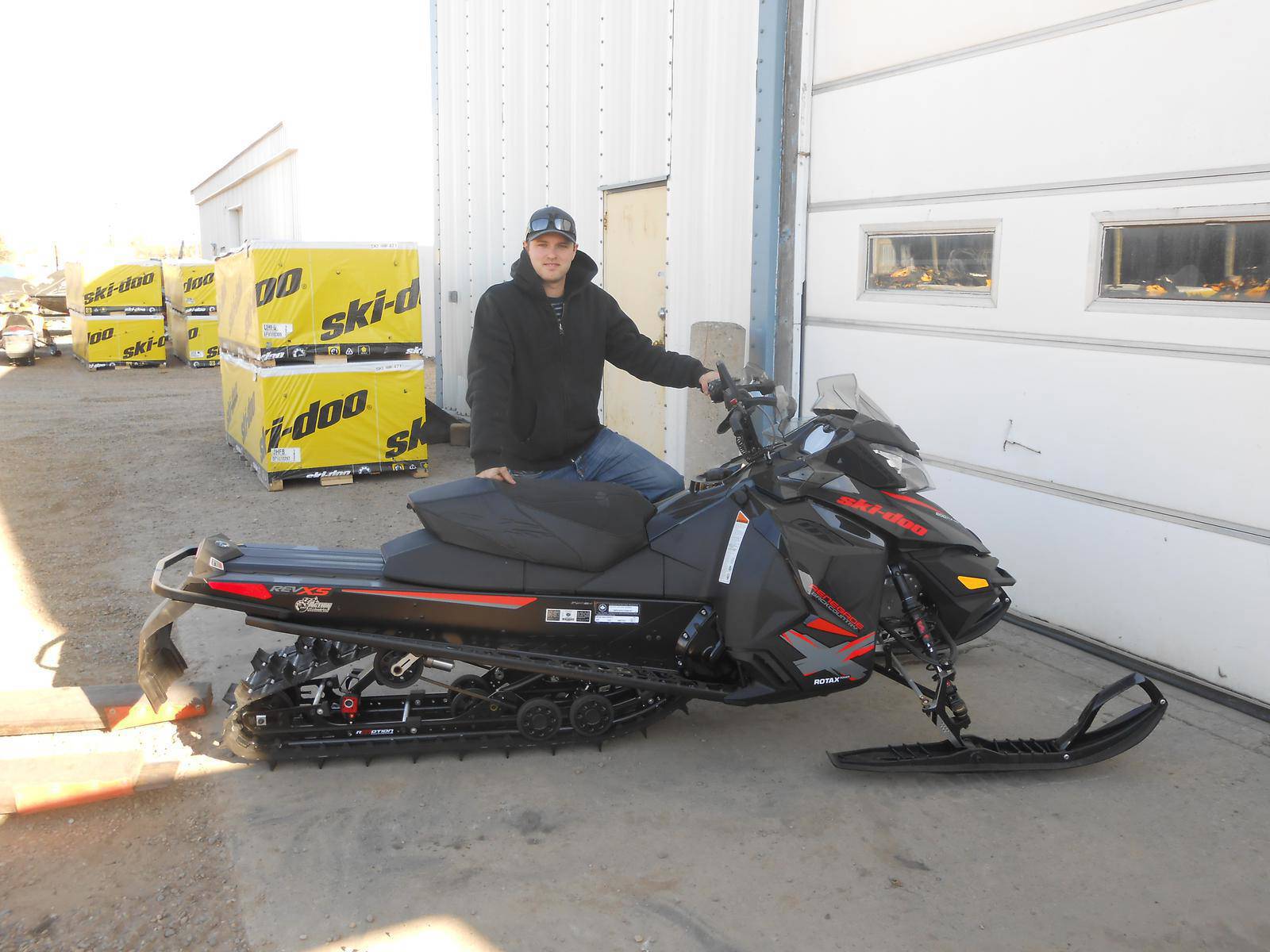 Man posing beside a black Ski-Doo snowmobile with red accents outside a dealership.
