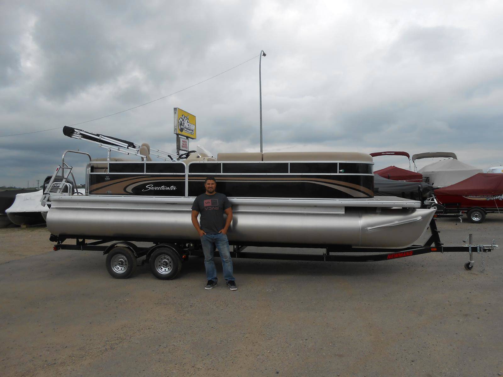 Man standing beside a black and tan Sweetwater pontoon boat on a trailer outside a dealership.