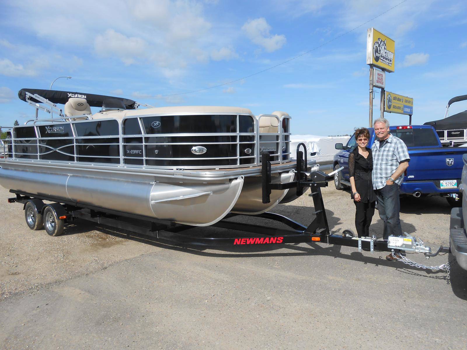 Man and woman standing beside a black and silver Xcursion pontoon boat on a trailer outside a dealership.